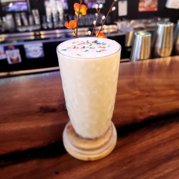 A tall glass of white milkshake with colorful sprinkles on top, on a wooden bar.