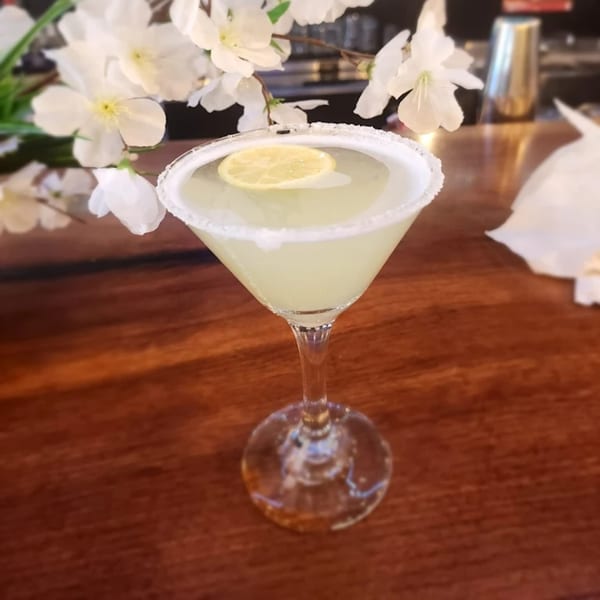 A light green cocktail with a sugared rim and lemon slice, next to white flowers on a wooden table.