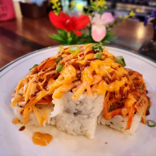 Sushi rolls topped with a creamy orange sauce, dark drizzle, and green onions on a white plate.