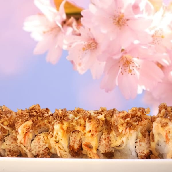 Sushi rolls topped with crispy fried onions and sauce, with pink cherry blossoms in the background.