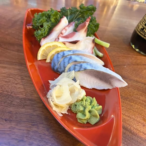 A red boat-shaped plate holds sushi, octopus, lemon, ginger, and wasabi on a wooden table.