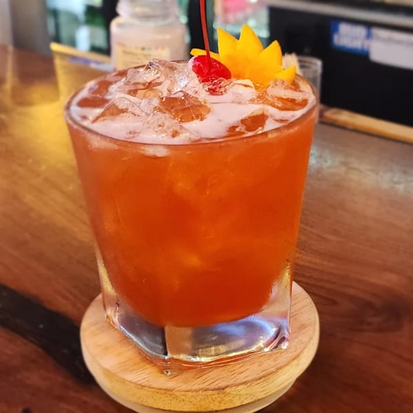A vibrant orange cocktail with ice, garnished with a cherry and mango slices, on a wooden bar.