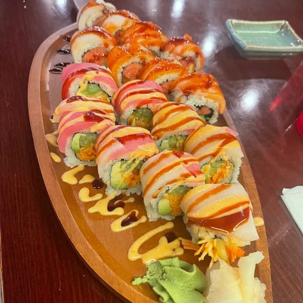 A wooden boat-shaped platter holds various sushi rolls, drizzled with sauces, alongside wasabi and ginger.