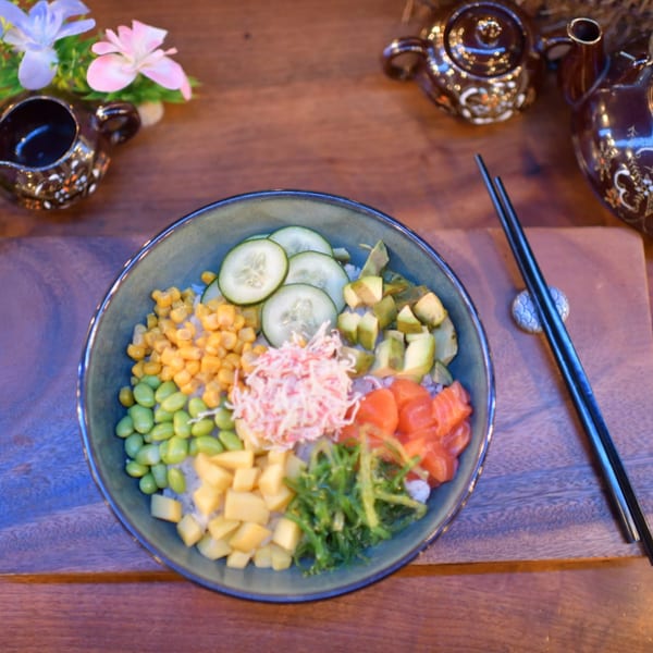 Colorful sushi poke bowl with salmon, crab, cucumber, corn, edamame, mango, and seaweed on a wooden table.