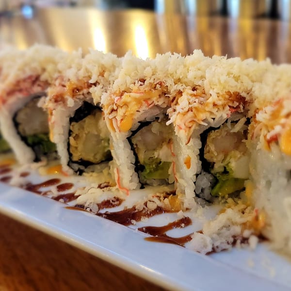 Close-up of sushi rolls topped with crispy tempura flakes and drizzled with sauce on a white plate.