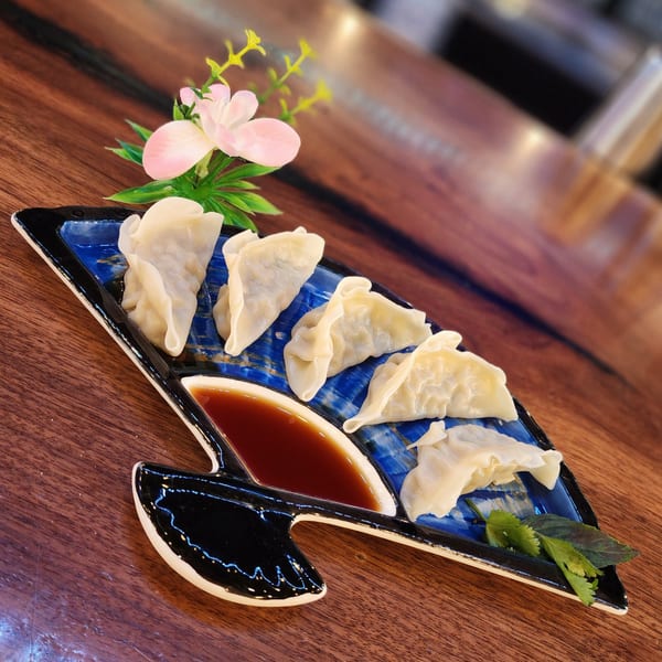 Six steamed dumplings with soy sauce and herbs on a blue and black fan-shaped plate.
